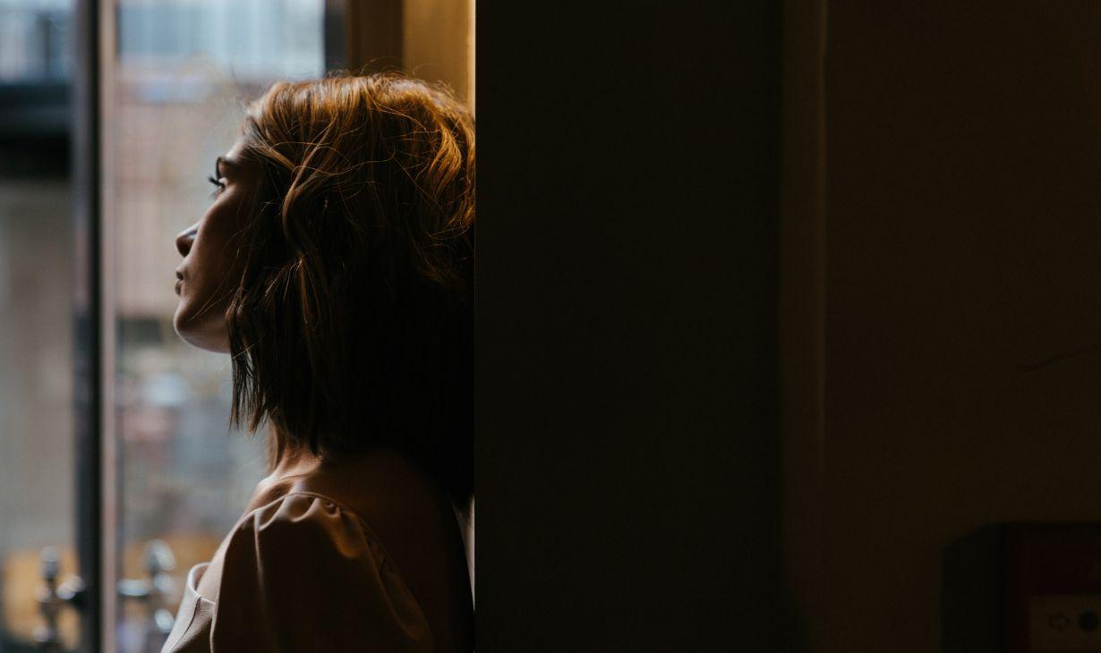 Une jeune femme, de profil, regarde par une fenêtre — pensive.